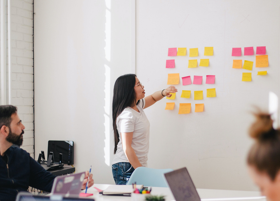 Woman points at white board with sticky notes in front of colleagues (1)
